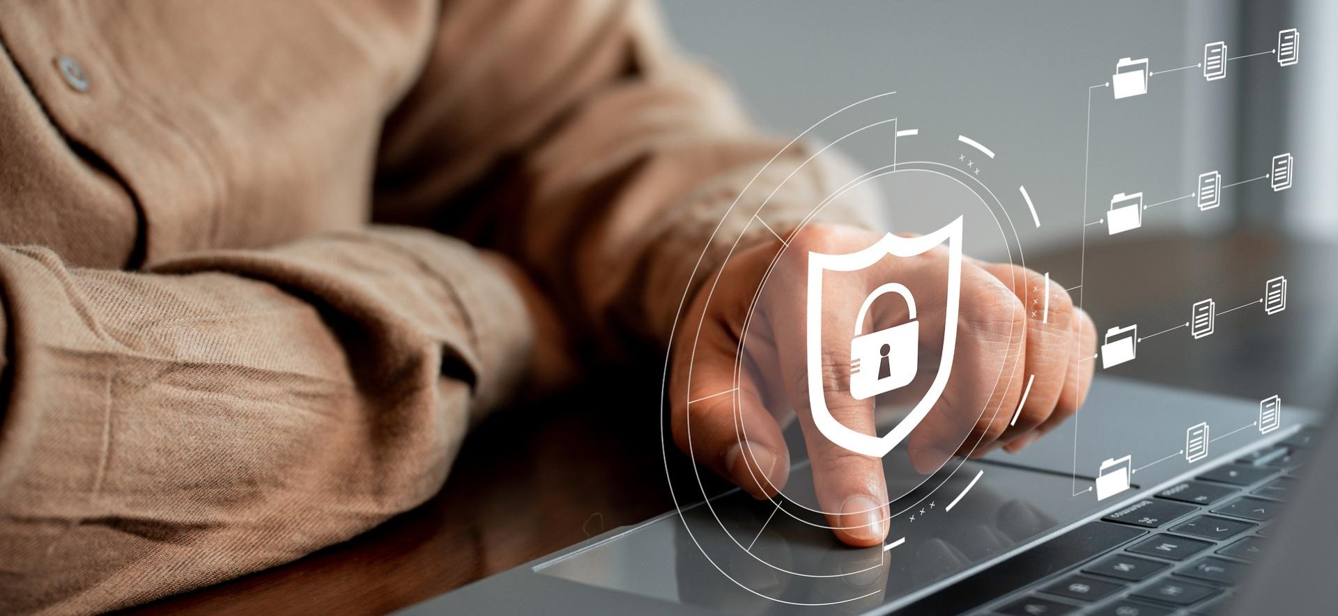 dłoń mężczyzny; palce na klawiaturze laptopa; na zdjęcie nałożony symbol tarczy i kłódki