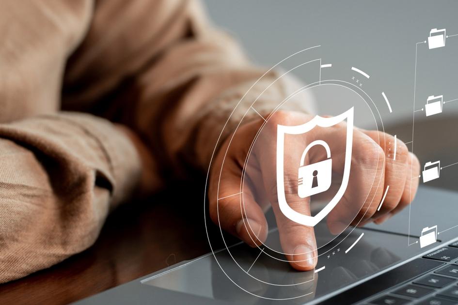 dłoń mężczyzny; palce na klawiaturze laptopa; na zdjęcie nałożony symbol tarczy i kłódki