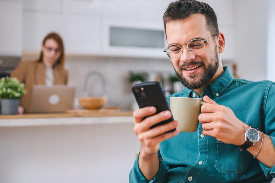Uśmiechnięty mężczyzna z kubkiem w dłoni i telefonem, coś klikający.